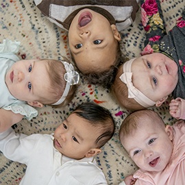 Five babies lay on the floor in a circle with their heads close together as they pose for a portrait.  They are each dressed casually and are smiling in this aerial view photo.