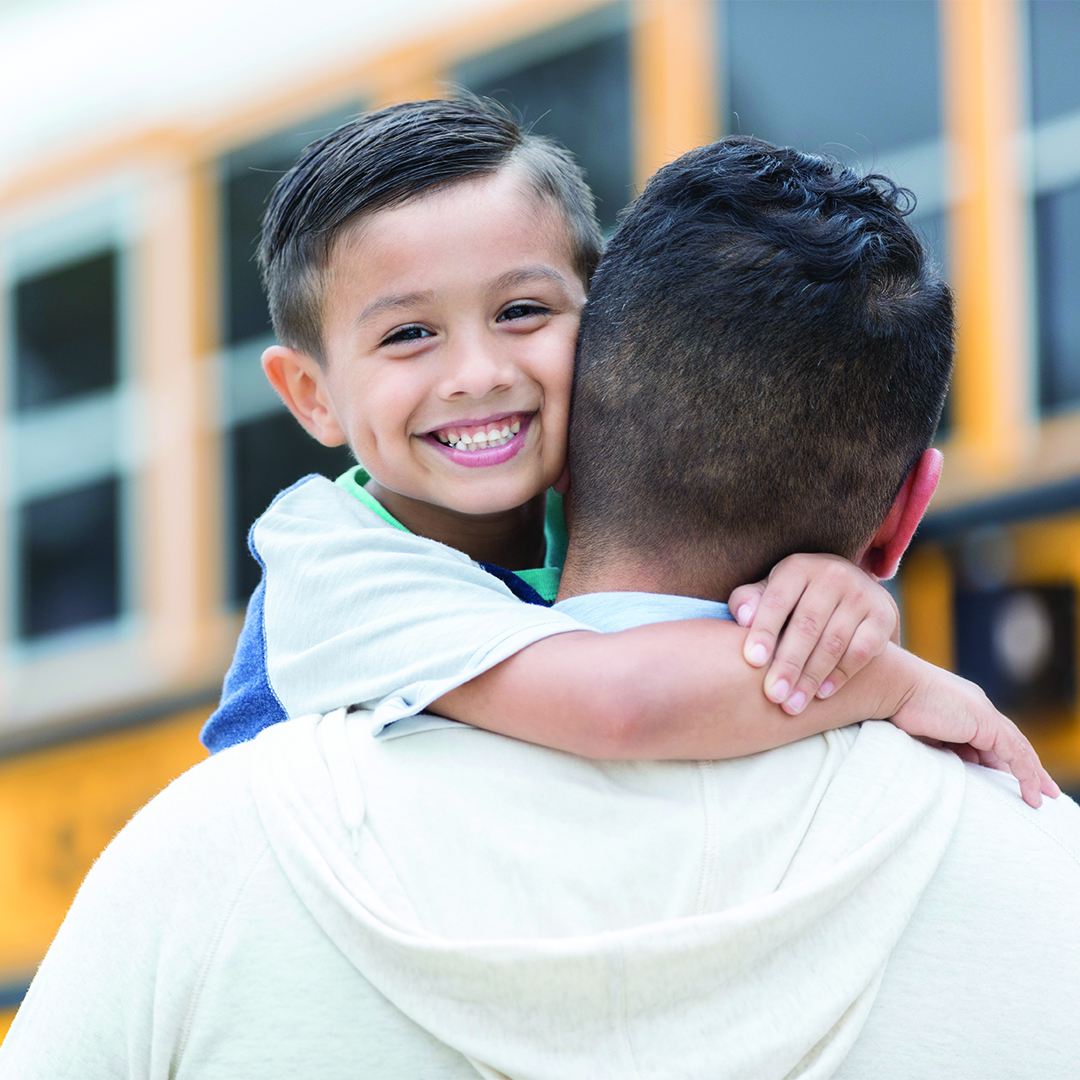 A confident little boy smiles for the camera as he embraces his father after the first day of school.  He has just ridden the school bus home.